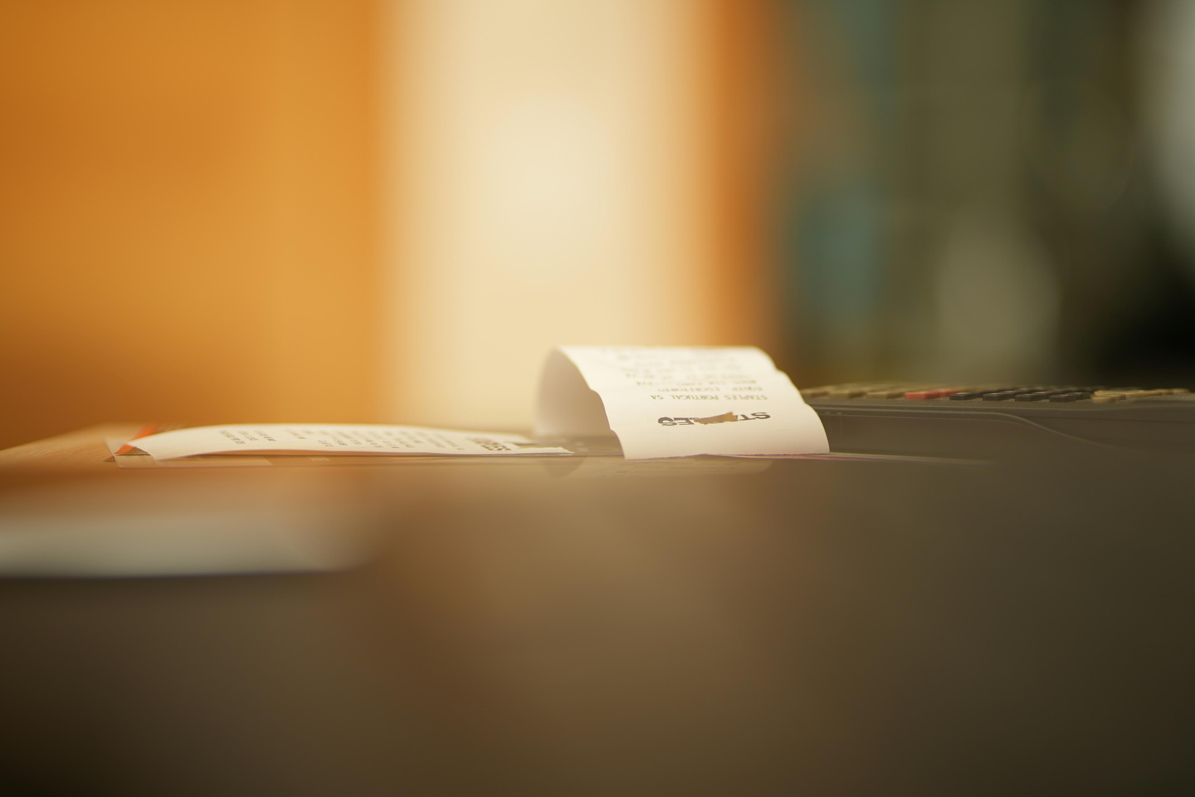 Paper invoices and documents stacked on a wooden desk by a sunlit window